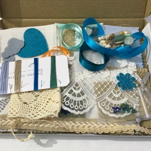 Peut inclure: Un kit de fournitures artisanales contenant une variété de matériaux, notamment des chutes de tissu, de la dentelle, du ruban, des boutons et de la laine. Le kit est présenté dans une boîte en carton et comprend une variété de couleurs, notamment le bleu, le blanc et le marron.