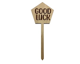 May include: A wooden sign with the words "GOOD LUCK" engraved on a pentagon-shaped top. The sign has a long, thin handle and is made of light-colored wood. The text is in a bold, sans-serif font.
