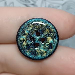 May include: A round, four-hole button featuring a glossy, swirling finish of deep teal, dark blue, and metallic gold tones. The surface has a glittery, reflective quality with an intricate, marbled pattern throughout the circular design.