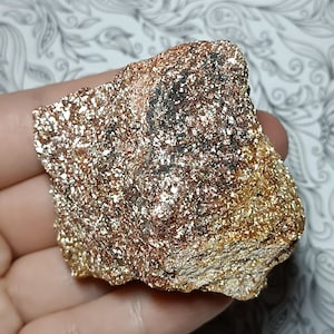 May include: A rough, irregularly shaped rock with a shimmering, metallic appearance. The surface is covered in small, reflective particles, creating a gold and bronze color. The rock is held in a hand, showcasing its texture and natural beauty.