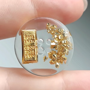 May include: A round, clear resin button featuring four center holes. Embedded inside is a small, rectangular gold-colored bar with the text "FINE GOLD 999.9" and a cluster of metallic gold-colored flakes arranged on the right side of the button.