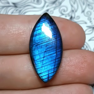 May include: A polished, marquise-shaped labradorite gemstone. The stone displays a vibrant blue iridescence with dark linear inclusions. The gemstone is held in a hand, showcasing its shape and color variations.