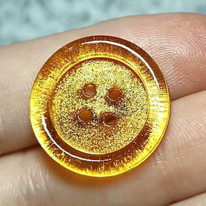 Puede incluir: Un botón redondo y translúcido de color ámbar con un centro texturizado y brillante, y un borde exterior liso y elevado. El botón tiene cuatro pequeños orificios espaciados uniformemente en el centro para su fijación, con un acabado brillante.
