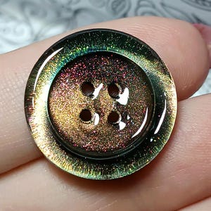 May include: A circular four-hole button featuring a glossy, iridescent finish that shifts between deep bronze, copper, and dark green tones. The surface has a fine, shimmering texture with a raised outer rim and a smooth, reflective appearance.