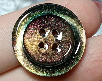 May include: A circular four-hole button featuring a glossy, iridescent finish that shifts between deep bronze, copper, and dark green tones. The surface has a fine, shimmering texture with a raised outer rim and a smooth, reflective appearance.