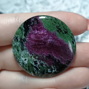 May include: A round, polished green and purple gemstone with black and white inclusions. The stone has a smooth, glossy finish.