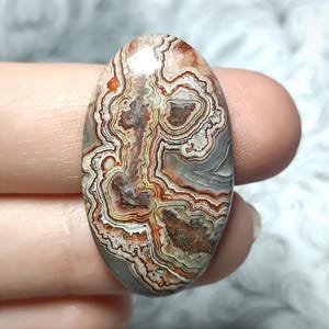 Puede incluir: Una gema ovalada con un patrón complejo de remolinos marrones, blancos, grises y naranjas. La piedra pulida se sostiene entre dos dedos, mostrando su belleza natural y variaciones de color únicas. La gema es una piedra natural.
