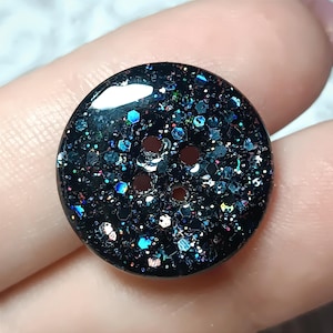 以下が含まれることがあります： 光沢のある黒いベースに、青、紫、銀の色合いで輝く多色の反射性六角形および円形のグリッター粒子が埋め込まれた、4つ穴の丸いボタン。留め具の表面全体にきらめきが広がっています。