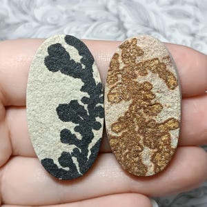 May include: Two oval-shaped stones. One stone has a beige background with a black, branching pattern. The other stone has a beige background with a brown, branching pattern. The stones are held in a hand.