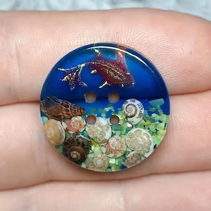 May include: A round, four-hole button featuring a clear resin design with a deep blue upper section containing a glittery, dark red fish silhouette. The lower section displays a collection of small, natural spiral seashells embedded in green.