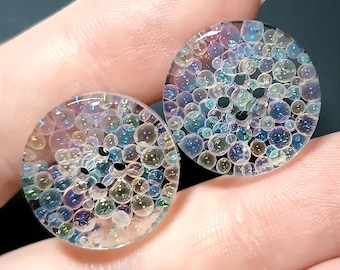 May include: Two circular, clear buttons featuring an embedded pattern of iridescent, multi-sized spheres that shimmer with blue, purple, and gold tones. Each button has four central holes for attachment, with a smooth, glossy surface finish.