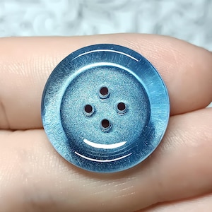 May include: A round, four-hole button with a shimmering, metallic blue finish. The button features a recessed center area and a slightly raised, translucent outer rim that catches the light, creating a distinct circular pattern on its surface.