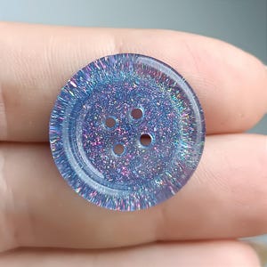 May include: A round, four-hole button featuring a translucent blue base embedded with fine, multi-colored iridescent glitter particles that create a shimmering, reflective effect across the entire surface of the circular fastener.