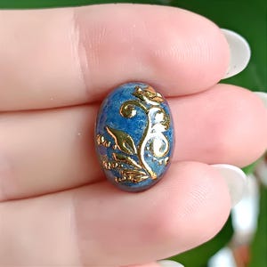 Könnte beinhalten: Ovale blauer Edelstein mit goldenem Blumenmuster. Das goldene Design zeigt Blätter und Wirbel. Der Edelstein wird zwischen Fingern gehalten, wodurch seine komplizierten Details und der Farbkontrast hervorgehoben werden.