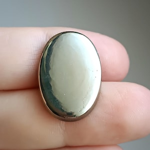 Pode incluir: Um botão metálico polido em forma oval com uma superfície refletora. O botão tem uma borda dourada e uma face lisa e brilhante. O botão está na mão de uma pessoa.