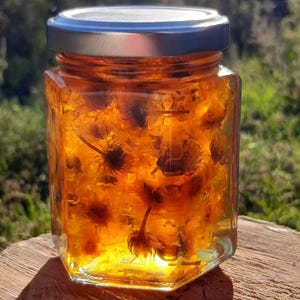 以下が含まれることがあります： 銀色の蓋が付いた透明な六角形のガラス瓶。オレンジ色の花が金色の液体に浸かっている。瓶は木製の表面に置かれ、背景はぼやけた緑色。