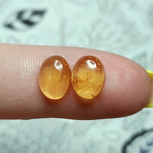 May include: Two oval-shaped, translucent amber gemstones with a golden hue and brown inclusions. The stones are smooth and polished.