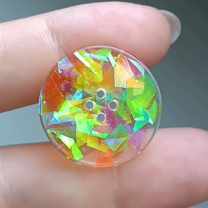 Puede incluir: Un botón redondo y transparente con incrustaciones de escamas de papel iridiscente multicolor en tonos verde, naranja y amarillo. El botón tiene una superficie lisa y brillante e incluye cuatro orificios centrales en forma cuadrada.