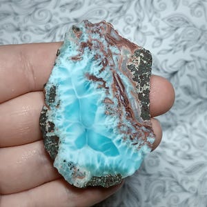 May include: A polished, irregular-shaped gemstone with a striking blue and white pattern, reminiscent of ocean waves. The stone has areas of brown and dark gray along the edges, adding contrast to the overall appearance. This unique piece is likely a decorative item or jewelry component.