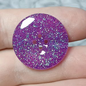 Op de afbeelding: Een ronde, platte knoop met een levendige paarse basis ingebed met fijne, reflecterende glitterdeeltjes die het licht vangen. De knoop heeft vier kleine, gelijkmatig verdeelde ronde gaten in het midden voor bevestiging, met een gladde afwerking.