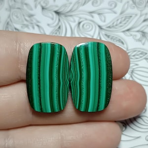 Puede incluir: Dos piedras de malaquita pulidas con esquinas redondeadas, que muestran un color verde vibrante con bandas paralelas de color verde oscuro. Las piedras se sostienen en una mano sobre un fondo estampado.