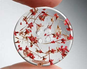 May include: A circular, clear resin button featuring embedded small, dried red and white flowers throughout its structure. The button has four central holes for attachment and a smooth, glossy finish, showcasing the delicate floral details.