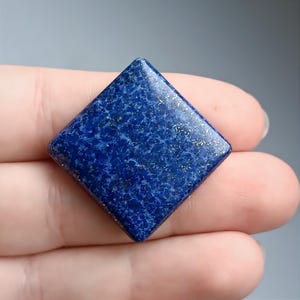 May include: A square, deep blue gemstone with gold flecks. The gemstone is held in a hand, showcasing its polished surface and unique texture. The stone's shape is a perfect square, and the color is a rich, saturated blue.