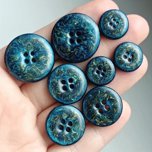 May include: A collection of seven round buttons in varying sizes, each featuring a deep blue base with swirling metallic gold and teal patterns. Every button has four central holes for attachment, showcasing a glossy, marbled finish throughout.