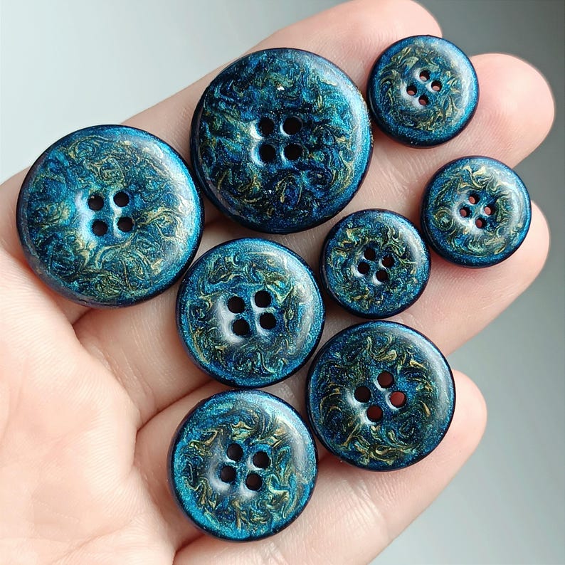 May include: A collection of seven round buttons in varying sizes, each featuring a deep blue base with swirling metallic gold and teal patterns. Every button has four central holes for attachment, showcasing a glossy, marbled finish throughout.