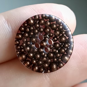 May include: A clear, circular button with a bronze-coloured interior filled with small, spherical beads. The button has four holes for sewing and a glossy finish. This unique button is suitable for various sewing and craft projects.
