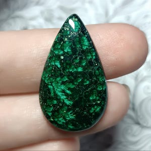 May include: A teardrop-shaped gemstone with a deep green color and a unique, fern-like pattern. The polished surface reflects light, highlighting the intricate details within the stone. The gem is held between fingers, showcasing its size and shape.
