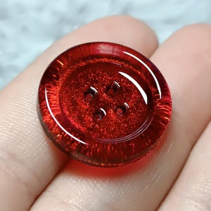 Puede incluir: Un botón redondo de color rojo translúcido con una textura brillante y centelleante en toda su superficie. El botón presenta un borde exterior elevado y estriado, y cuatro pequeños orificios equidistantes en el centro con acabado brillante.