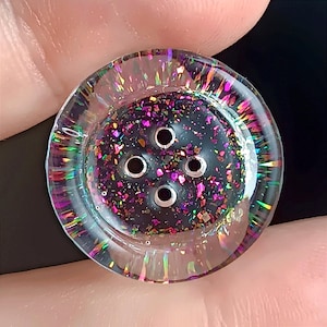 Puede incluir: Un botón redondo de cuatro agujeros con un centro oscuro lleno de escamas de purpurina iridiscente multicolor. El borde exterior está hecho de material transparente que muestra un vibrante efecto de refracción de luz tipo arcoíris.