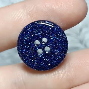 May include: A round, dark blue button with a glossy finish and embedded multi-colored glitter particles. The button features four centered holes arranged in a square pattern, providing a functional design for various sewing or craft projects.