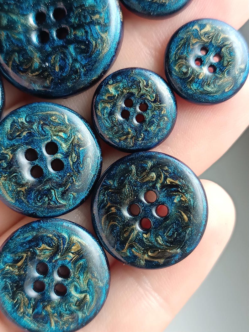 May include: A collection of round buttons featuring a deep blue and metallic gold swirling marbled pattern. Each button has a smooth, glossy finish and four central holes for attachment, showcasing a vibrant, reflective, and glittery texture.