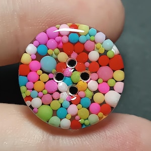 Op de afbeelding: Een ronde, doorzichtige knoop gevuld met een dichte opstelling van kleine, veelkleurige bolvormige kralen in tinten roze, blauw, rood, geel, oranje, groen en wit. De knoop heeft vier centrale gaten voor bevestiging en een glad oppervlak.