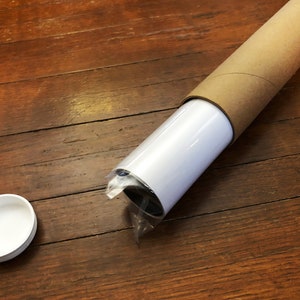 Puede incluir: Un p&oacute;ster o impresi&oacute;n enrollado est&aacute; asegurado en un tubo de cart&oacute;n marr&oacute;n. El tubo est&aacute; sobre una superficie de madera. Tambi&eacute;n se ve una tapa de pl&aacute;stico blanca en la imagen.