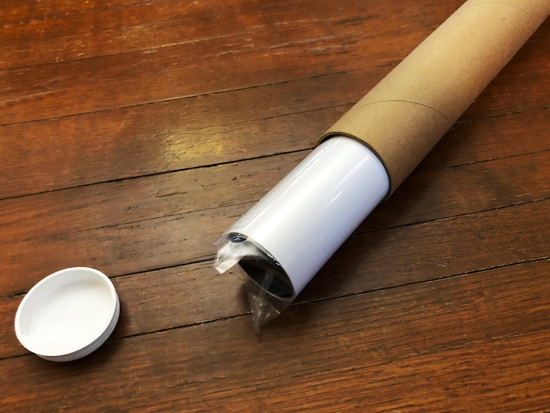 Puede incluir: Un p&oacute;ster o impresi&oacute;n enrollado est&aacute; asegurado en un tubo de cart&oacute;n marr&oacute;n. El tubo est&aacute; sobre una superficie de madera. Tambi&eacute;n se ve una tapa de pl&aacute;stico blanca en la imagen.