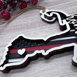 May include: Wooden ornament shaped like the state of Massachusetts with a red, white, and black thin red line flag design. The ornament has a small white heart in the center and the word "Home" is written on the side.
