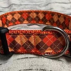 May include: A dog collar with a repeating argyle pattern in shades of orange, brown, and tan. The collar has a black plastic buckle and a silver metal D-ring for leash attachment.