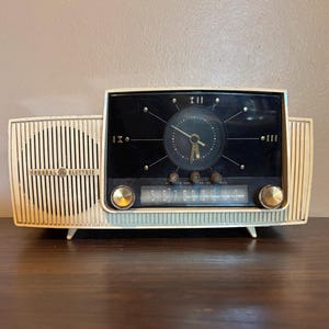 May include: A cream-colored General Electric radio with an integrated analog clock. The device features a vertical-slat speaker grille, a black clock face with gold-toned hands and markers, and a horizontal radio dial with three control knobs.