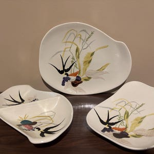 May include: Three white ceramic serving dishes with a mid-century modern design. Each dish features a unique shape and is decorated with a botanical motif in shades of green, yellow, black, and purple.