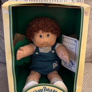May include: A Cabbage Patch Kids doll in a blue and white outfit, with brown curly hair and freckles. The doll is sitting in a box with a green background and the Cabbage Patch Kids logo. The box also includes a birth certificate and adoption papers.