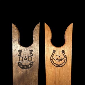May include: Two wooden boot jacks with a horseshoe design. One boot jack is dark brown and has the word "DAD" engraved inside the horseshoe. The other boot jack is light brown and has a cowboy hat engraved inside the horseshoe.