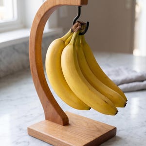 May include: A wooden banana stand featuring a curved arm and a rectangular base. A black metal hook is attached to the underside of the arm, holding a bunch of five yellow bananas. The stand is constructed from light brown wood with a visible grain.