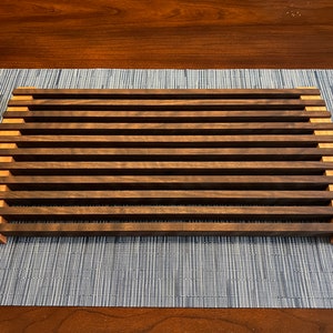 Puede incluir: Un estante de enfriamiento de madera con un marco marrón claro y listones marrones oscuros. El estante está hecho de madera y tiene una forma rectangular.