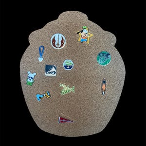 May include: A corkboard with a variety of pins, including a Mickey Mouse pin, a Star Wars pin, a pin with a fishbowl, and a pin with a pencil. The corkboard is shaped like a heart.