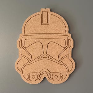 Puede incluir: Una pieza de arte mural de madera de color marrón claro que representa un casco de Clone Trooper. La obra de arte está sobre un fondo gris. El diseño del casco incluye la visera, la boca y otros detalles.