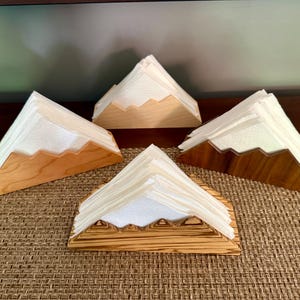 Könnte beinhalten: Vier Serviettenhalter aus Holz, jeweils mit einem Bergspitzen-Design, die weiße Servietten halten. Die Halter variieren im Holzton, von hell- bis dunkelbraun. Die Halter sind auf einer gewebten, braunen Oberfläche angeordnet.