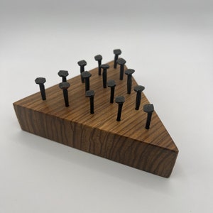 Puede incluir: Un bloque de madera triangular con un acabado de madera oscura. El bloque tiene 15 clavos de metal negro martillados en él en un patrón triangular.